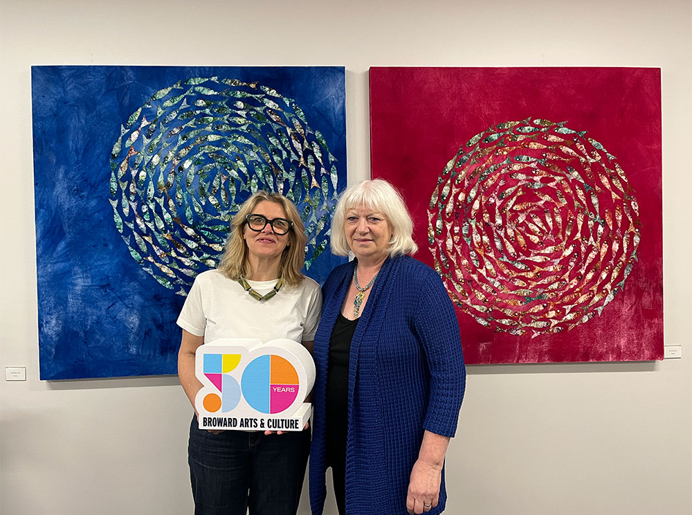 Anna Paola Cibin and Louise Irvine Splash! Exhibition at WMODA, standing in front of two velvet paintings. They are holding the Broward County Cultural Division 50 Year logo.
